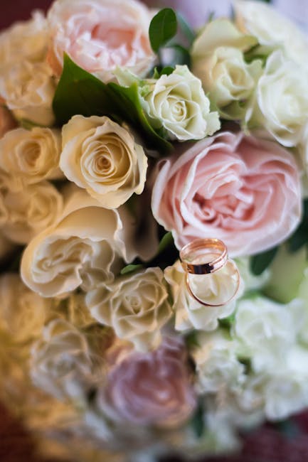 A close-up of a wedding bouquet featuring soft pink and creamy white roses, arranged with lush green leaves and wrapped in elegant pastel-colored paper. The flowers exhibit fresh, delicate petals with