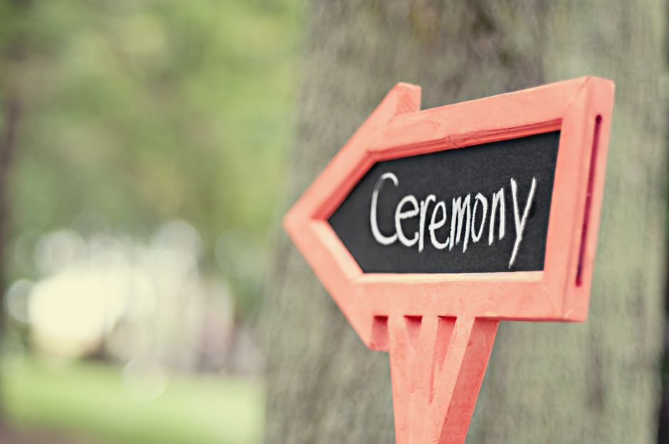 A close-up of a pink wooden directional sign with a black chalkboard surface, featuring the word 'Ceremony' written in white chalk. The sign is attached to a tree trunk outdoors, with a blurred backgr