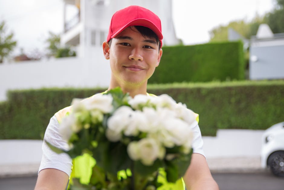 A male flower delivery courier wearing a red cap and a high-visibility yellow vest is holding a bouquet of fresh white flowers, possibly roses or peonies, with green leaves. The bouquet is wrapped in 